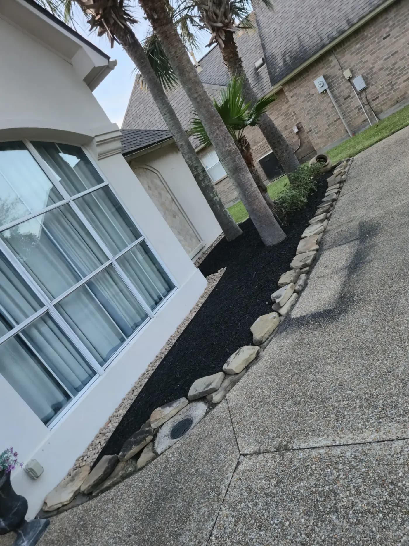 Fresh black mulch bed bordered by light flagstones along a white house and aggregate driveway.