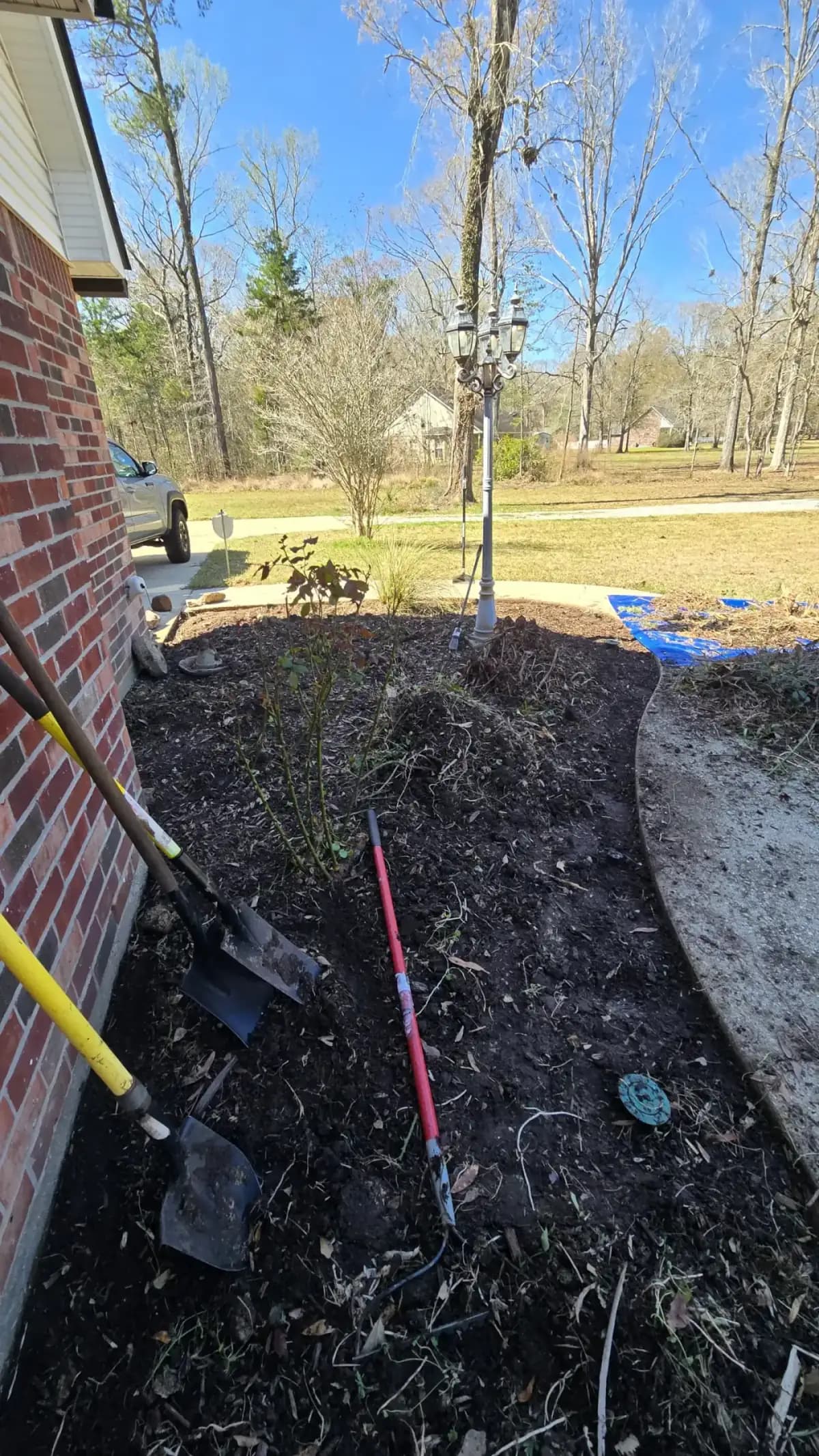 Shovels and cultivator in a freshly turned flower bed along a brick house under blue sky.