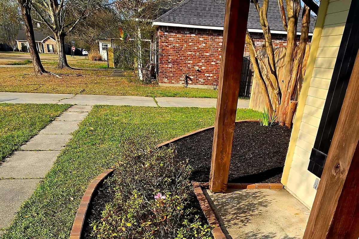 New landscape bed in Walker, LA with brick edging, fresh black mulch, and a young shrub beside a porch post.