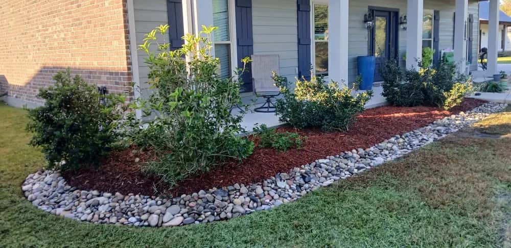 New landscape bed in Walker, LA with river rock edging, fresh red mulch, and shrubs along a front porch.